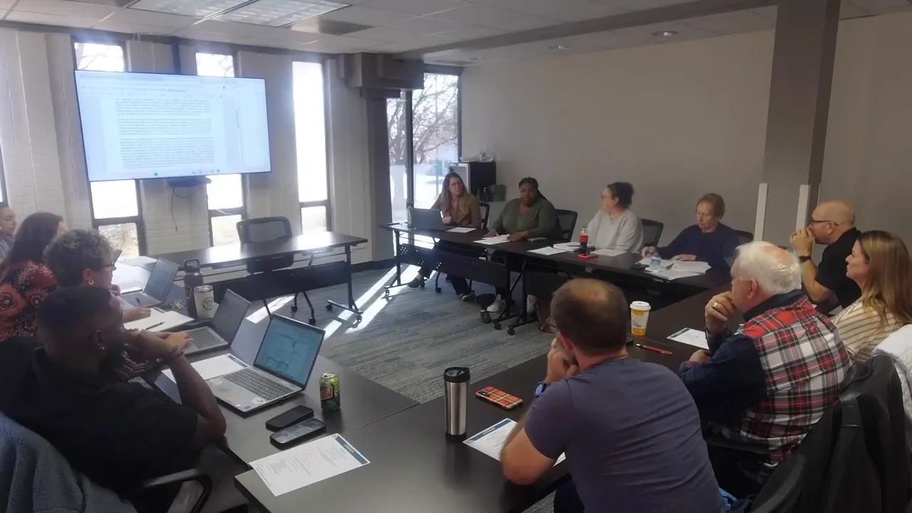 Clear overhead view of Longmont Housing Authority board members around a table with a wall-mounted screen showing agenda text.