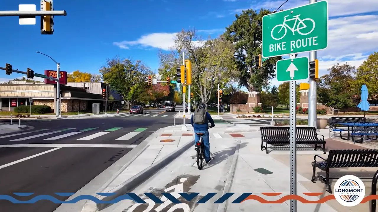 Cyclist riding in a separated bike lane toward a corner island and green bike crosswalk with a 'Bike Route' sign visible