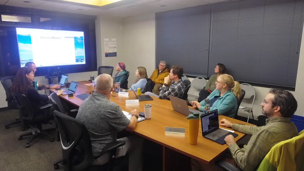 Meeting-room wide shot with advisory board members at a table and a clear view of the large screen showing the ServiceWorks work-order interface.