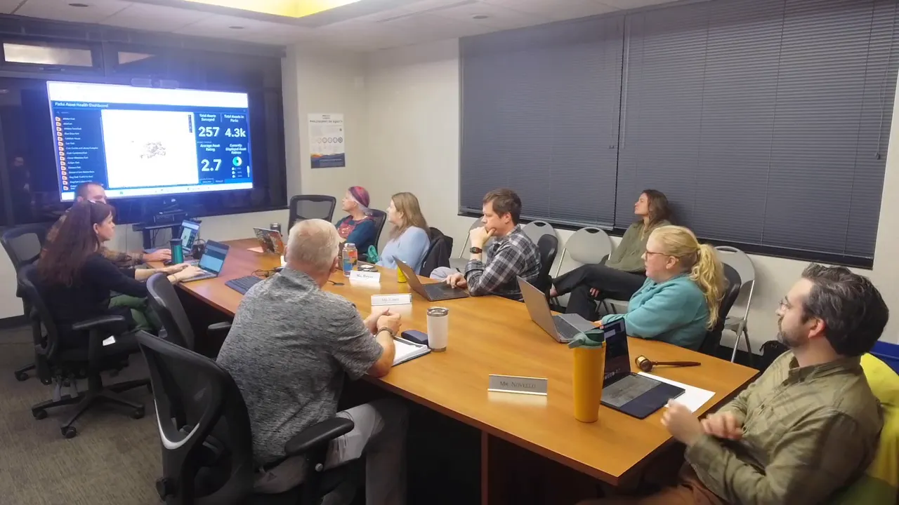 Wide shot of a parks advisory board around a table watching a TV that displays the Total Park Health dashboard with an average asset rating.