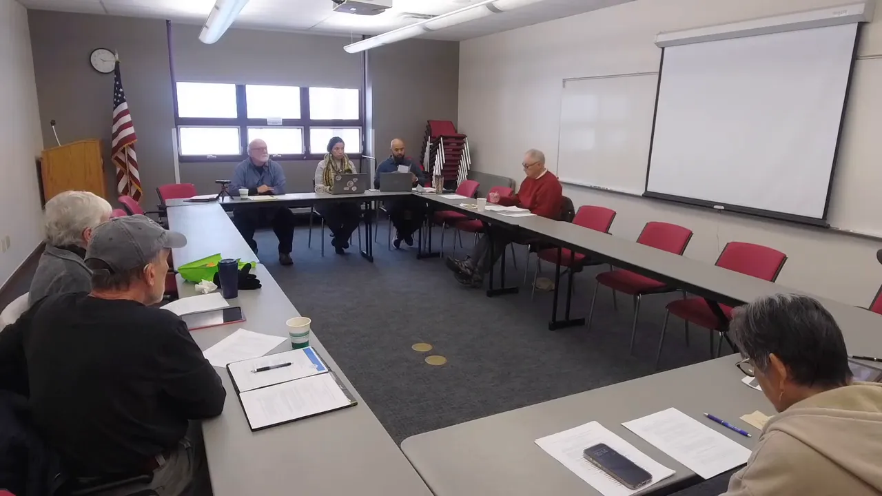 Clear wide-angle photo of the senior advisory board meeting showing members around a U-shaped table, documents and laptops visible, with a projector screen in the background.