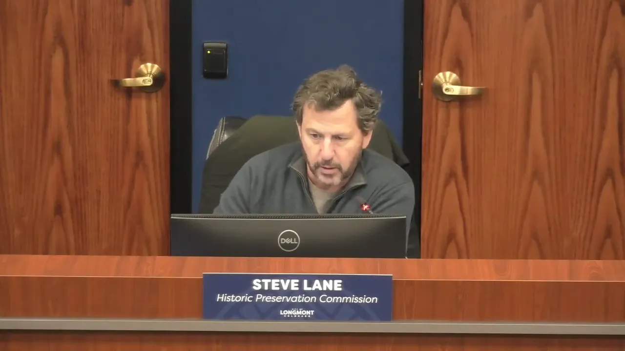 Frontal view of a commission member at the dais behind a monitor and a commission placard; well-lit, clear, and focused.