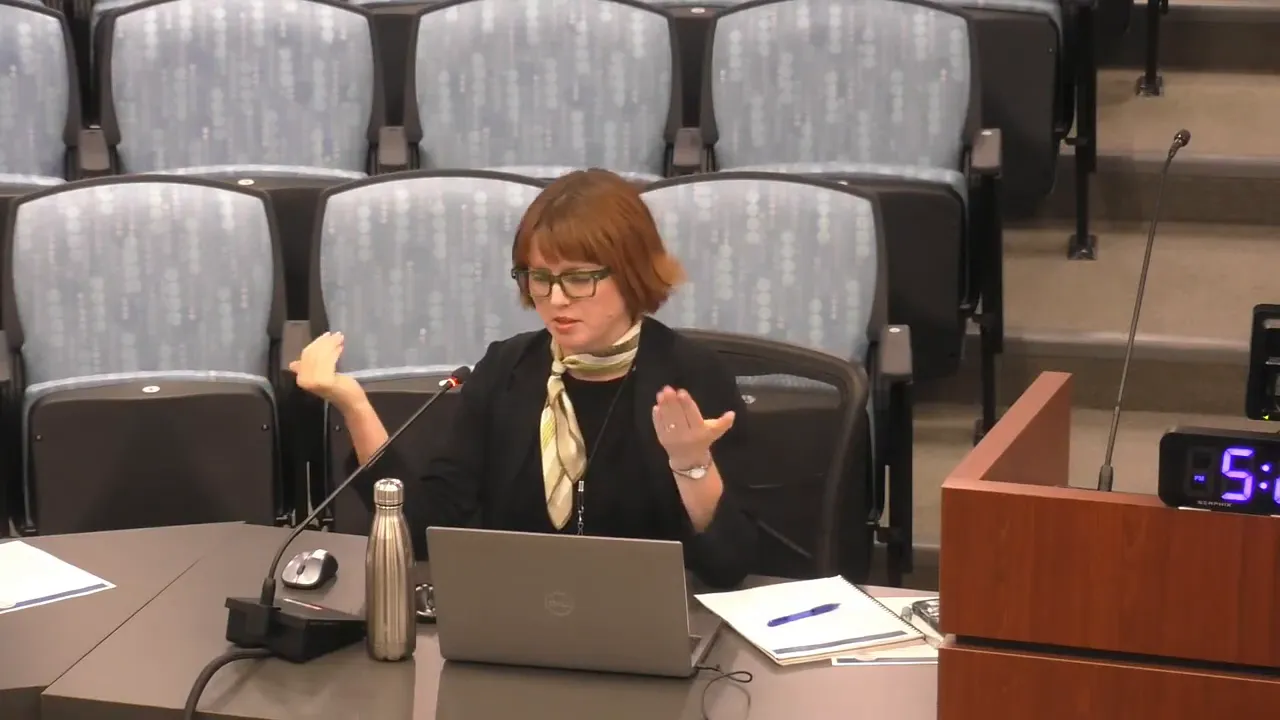 Commissioner at dais gesturing with both hands while speaking; laptop, notes, and water bottle visible on the table in front of her, empty audience seats in background.