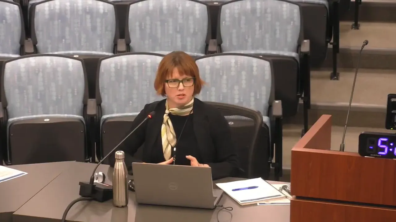 Mid-shot of a commissioner at the dais speaking into a microphone with hands gesturing, laptop and documents on the table and auditorium seats in the background.