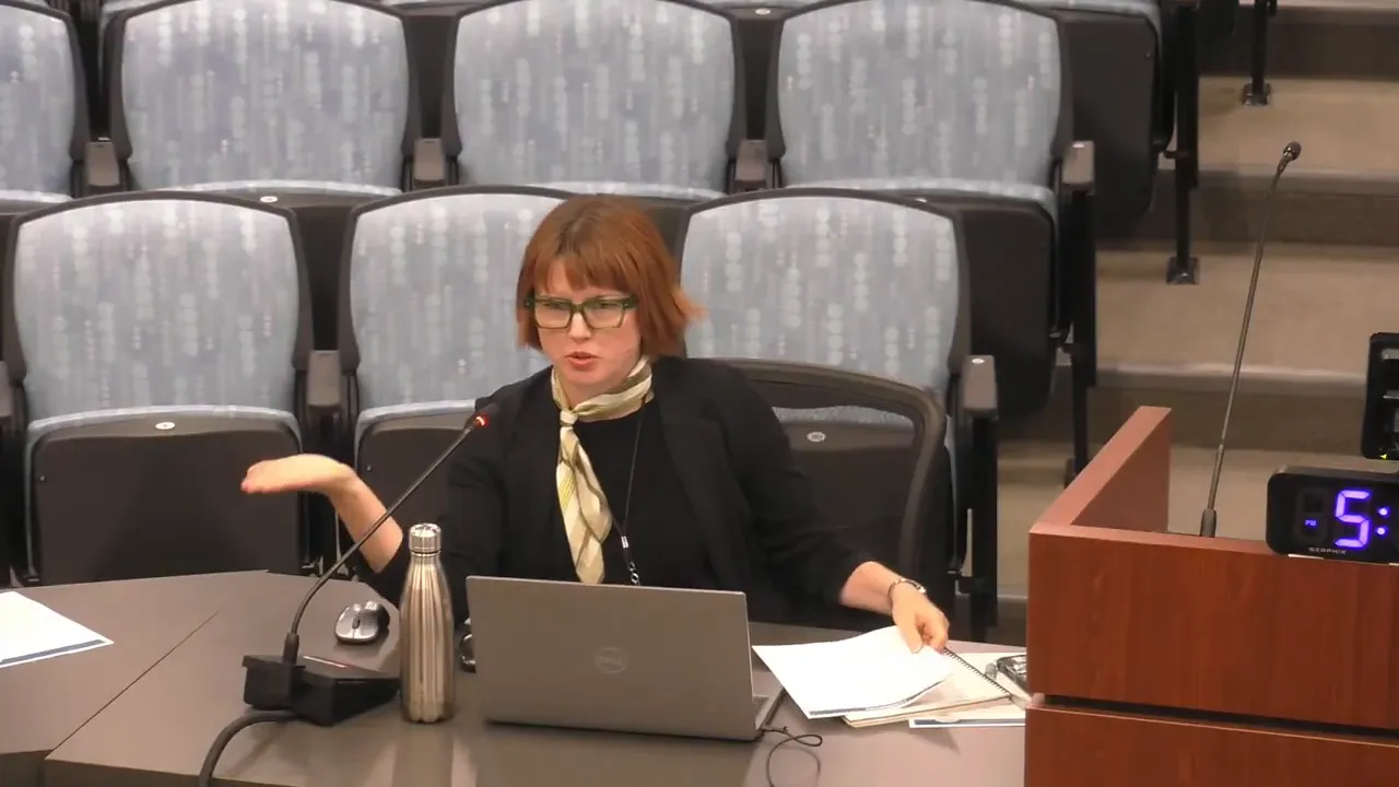 Clear, high-quality image of a commission member at the dais gesturing with one hand and holding meeting papers with the other, laptop and microphone visible.
