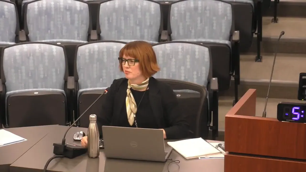 Commissioner at the dais speaking into a microphone with a laptop and documents visible; empty auditorium seats in the background.