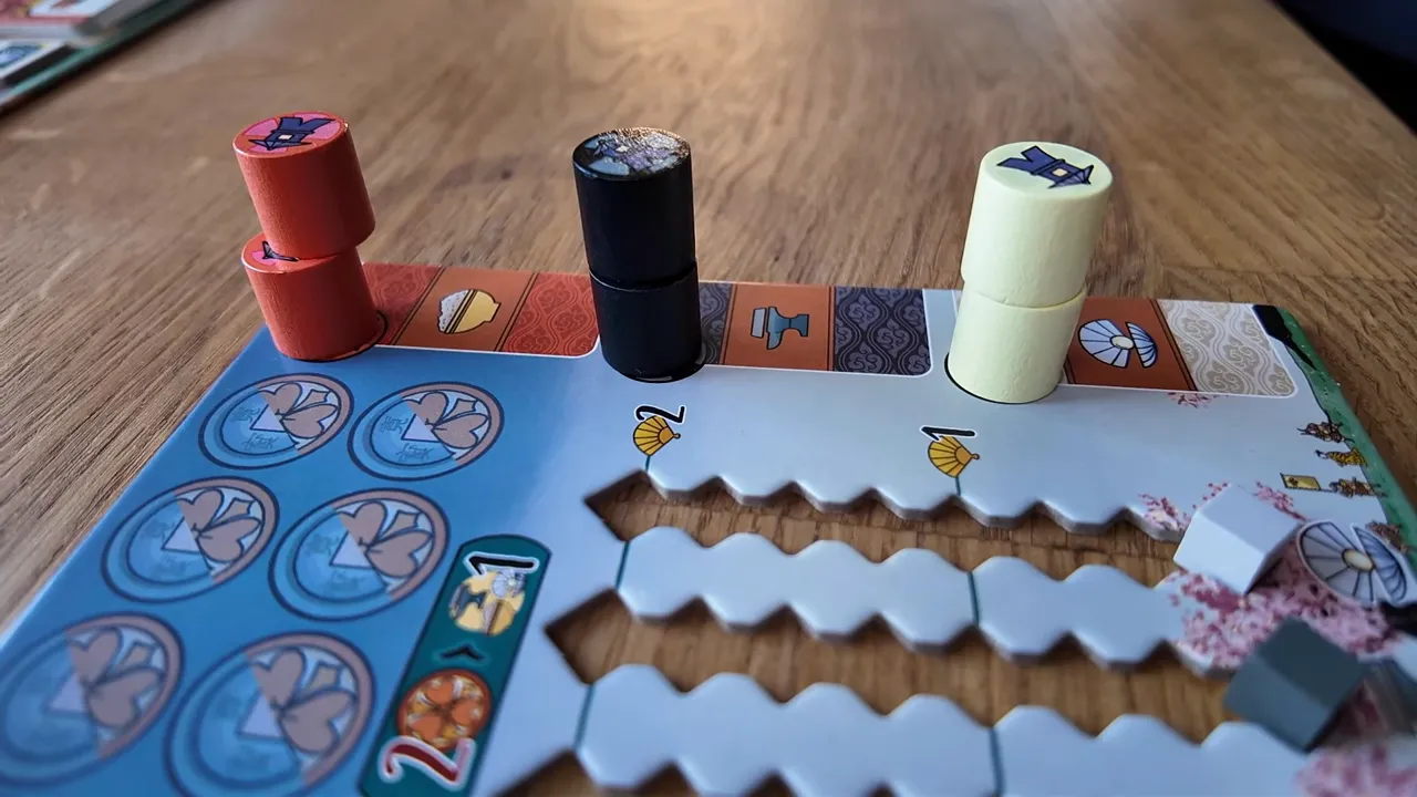 Makroaufnahme eines Spielertableaus mit Holzmarkern (rot, schwarz, gelb) auf Holztisch; Detail des Komponenten-Designs sichtbar.