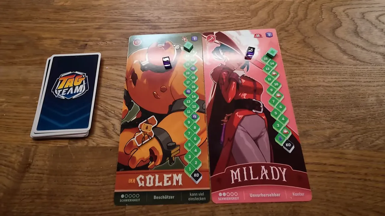 Nahaufnahme der Tag Team Charakter‑Boards 'Der Golem' und 'Milady' neben dem Kartenstapel auf Holztisch