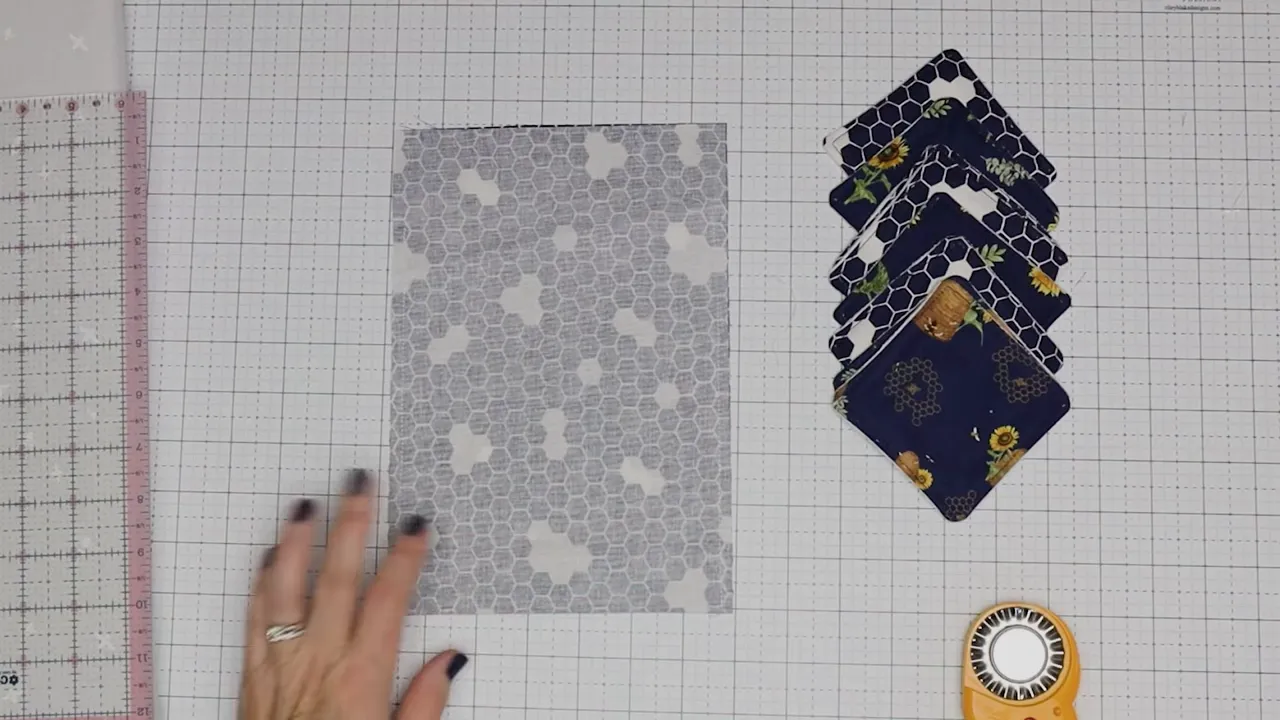 Top-down view of a prepared fabric rectangle, a neat stack of finished reusable makeup pads, and a rotary cutter on a gridded cutting mat.