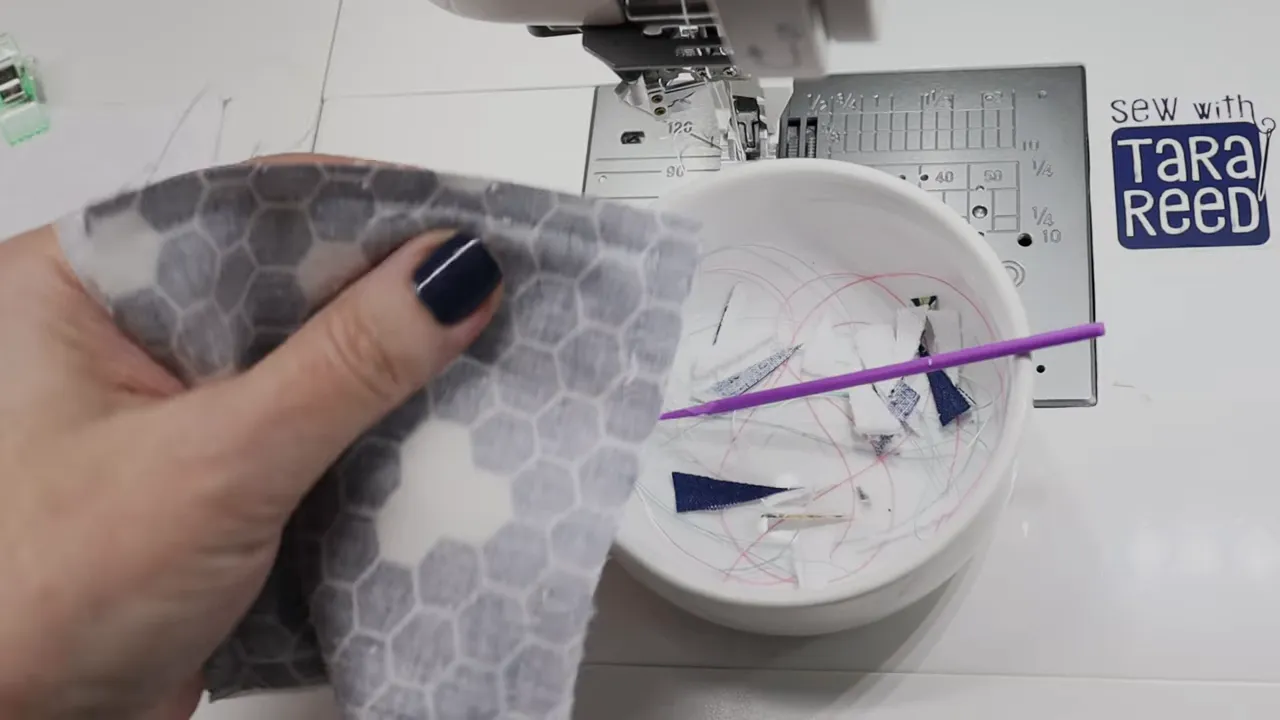 Hand holding a fabric pad near a sewing machine throat plate with a bowl of thread trimmings nearby.