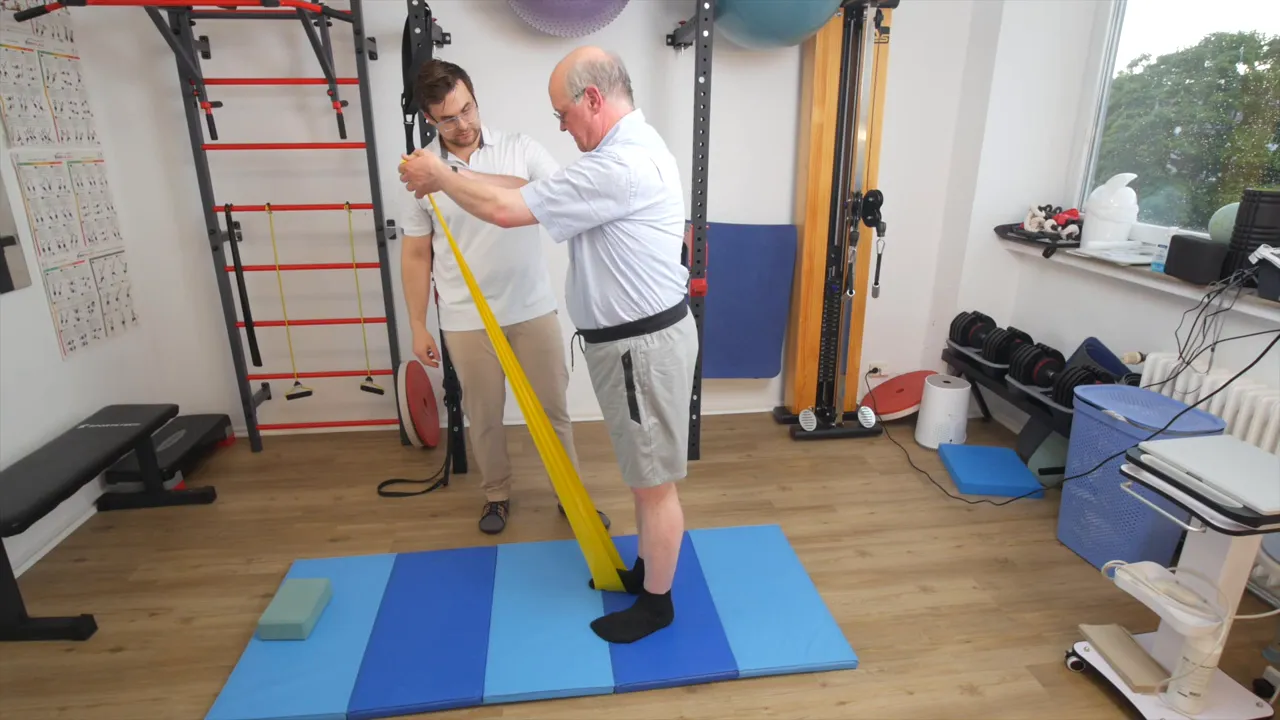 Volle Übungsszene: Patient steht auf Matte mit Theraband unter dem Fuß und führt einen kontrollierten Armzug aus, Therapeut beobachtet