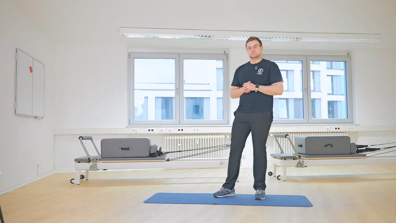 Physiotherapeut in heller Praxis steht auf einer Gymnastikmatte vor Trainingsgeräten und bereitet eine Übung vor