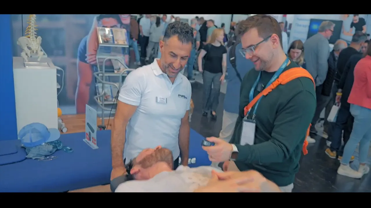 Drei Personen an einem Messestand demonstrieren Mobilisation an der Manuthera 242; ein Patient liegt auf der Liege