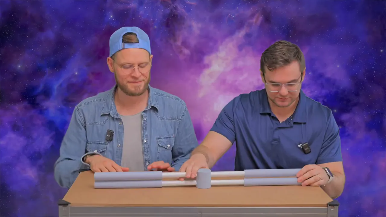 Zwei Moderatoren sitzen am Tisch und zeigen den Schaumstoff‑Haltungstrainer mit deutlich sichtbarem zentralem Verbinder und Polstern.