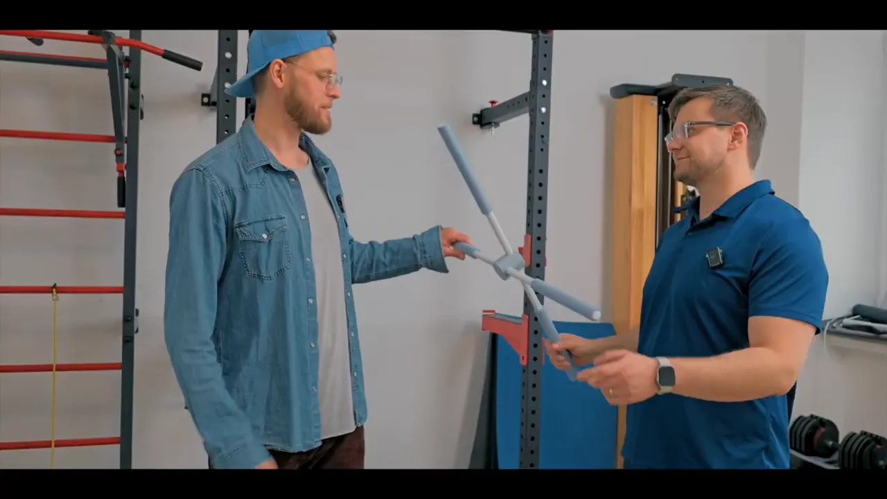 Zwei Trainer halten den gepolsterten Dreipunkt‑Haltungstrainer frontal, zentraler Verbinder und drei Stangen klar sichtbar.