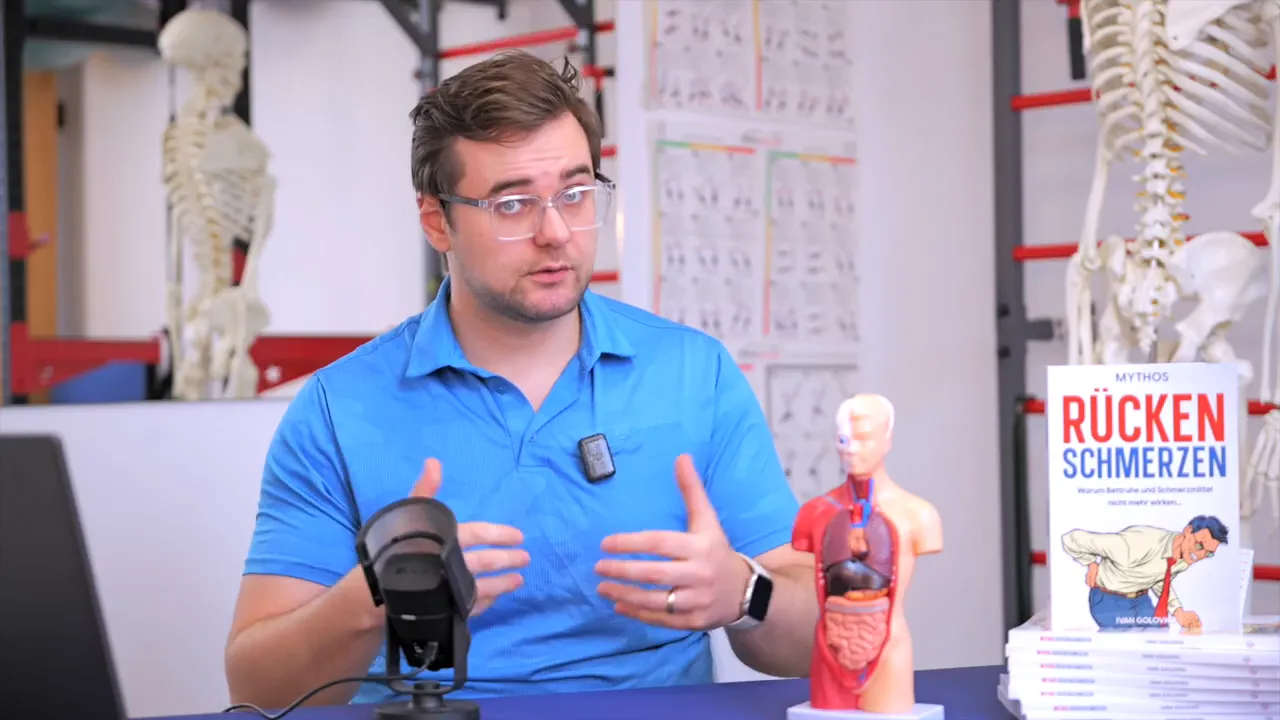 Physiotherapeut erklärt mit Handgesten neben einem anatomischen Torso‑Modell und dem Fachbuch 'Rückenschmerzen'.
