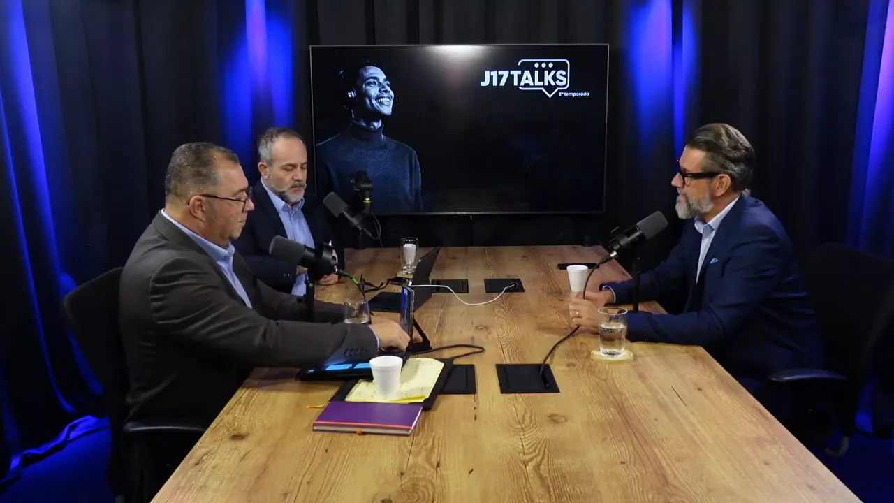 Vista ampla da mesa com três participantes conversando em estúdio e tela ao fundo