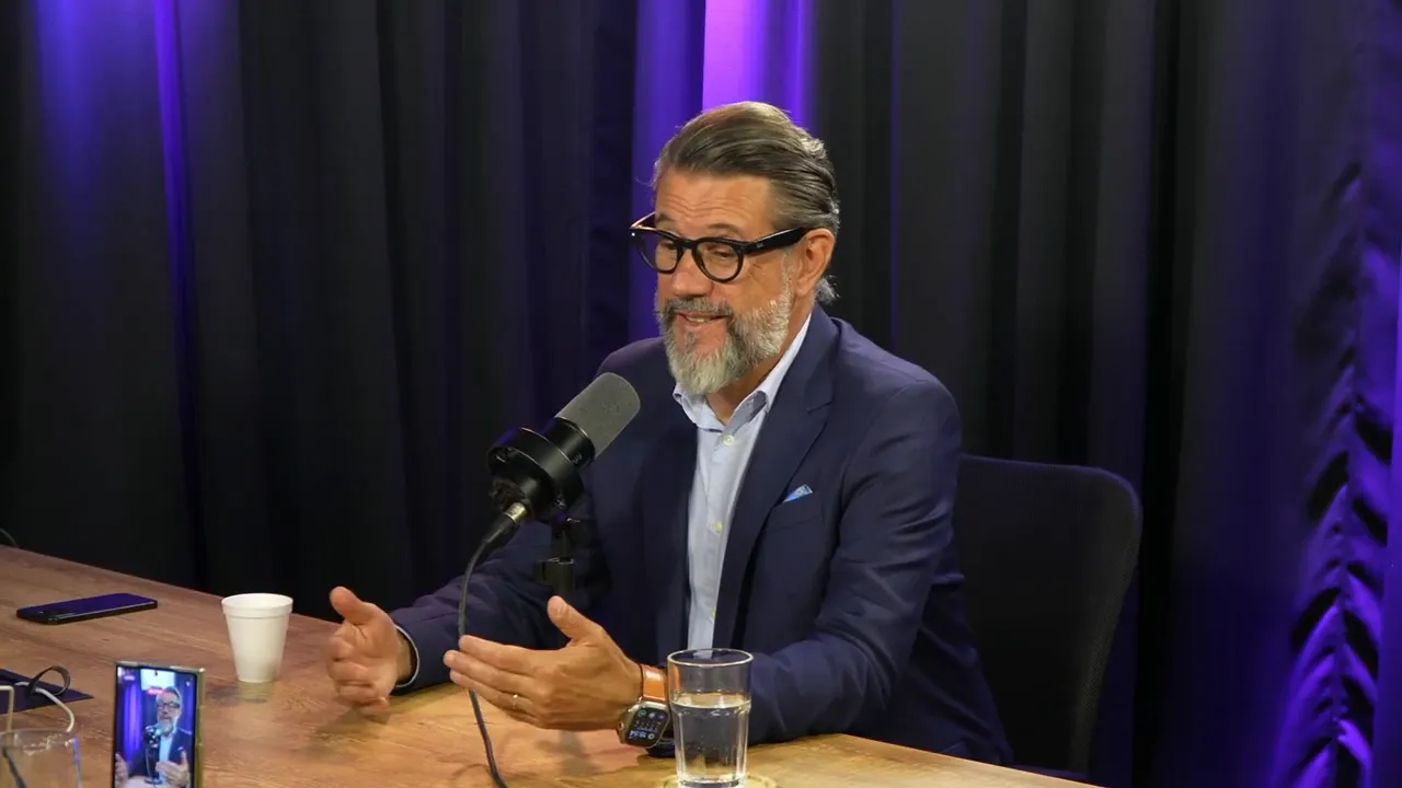 Convidado em estúdio com as mãos estendidas explicando, microfone e copo sobre a mesa