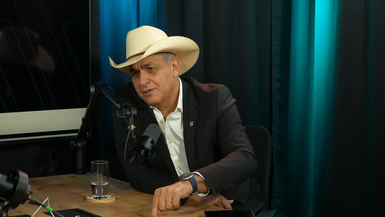 Convidado de chapéu claro falando ao microfone e gesticulando com a mão sobre a mesa