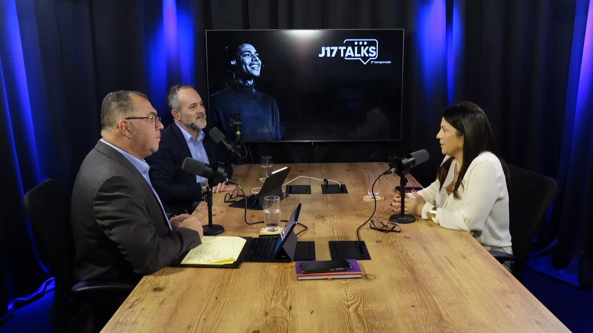 Mesa de estúdio com três participantes em traje profissional conversando em podcast