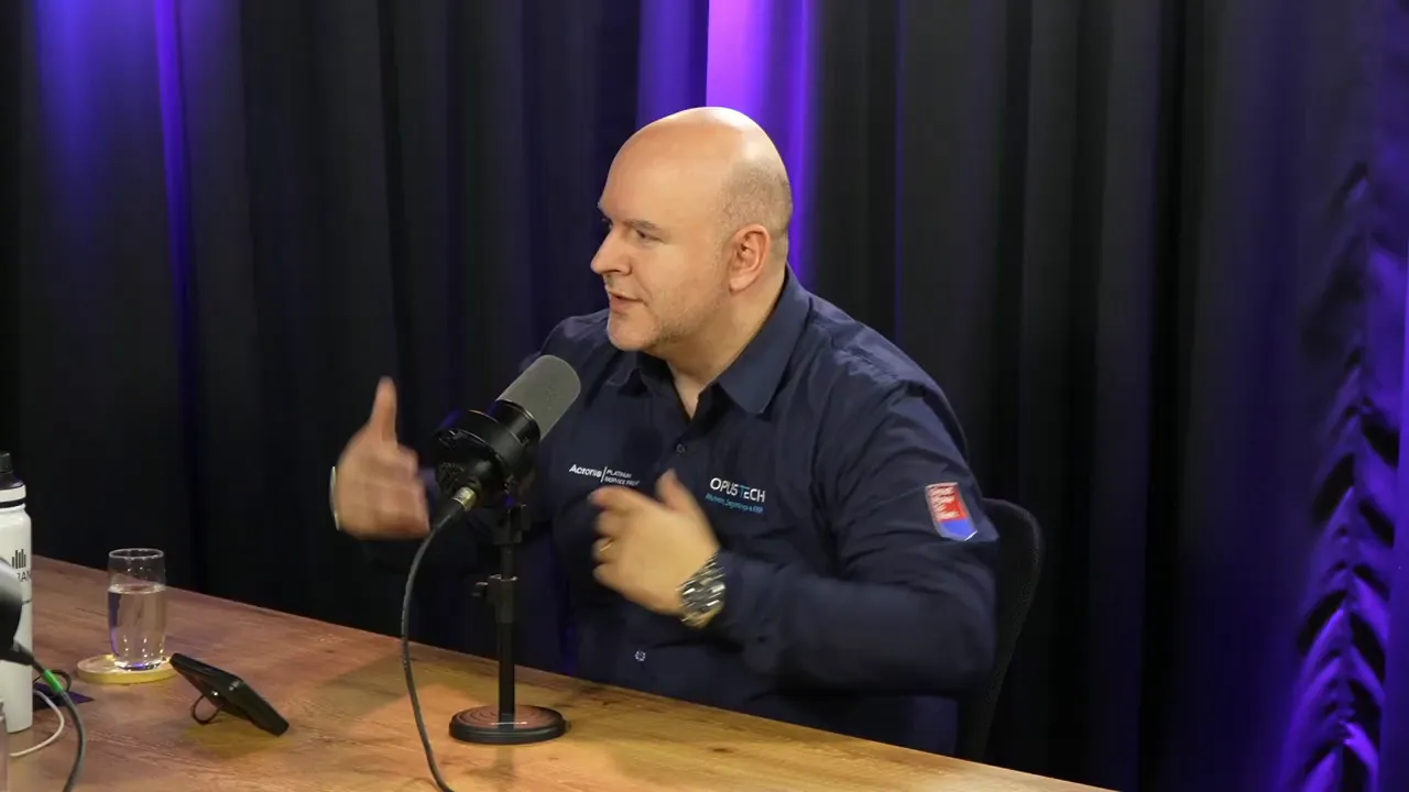 Pessoa gesticulando enquanto fala em podcast, microfone central e cortina com iluminação roxa ao fundo