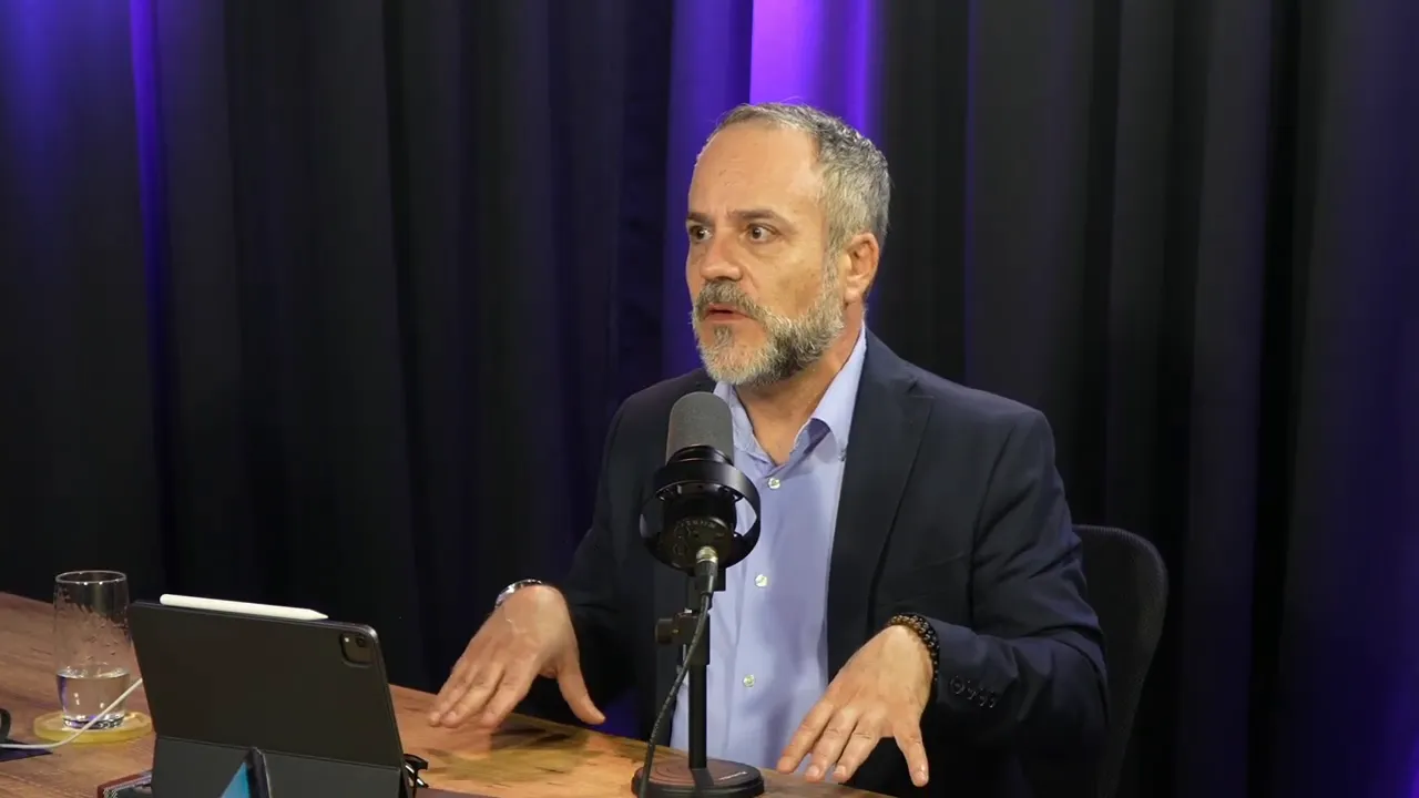 Apresentador em estúdio olhando para o lado, gesticulando com as mãos diante de um microfone durante uma explicação