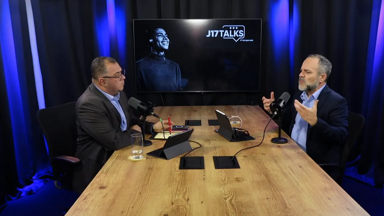 dois participantes em debate à mesa com microfones, gesto expressivo e tela ao fundo mostrando ambiente de talk
