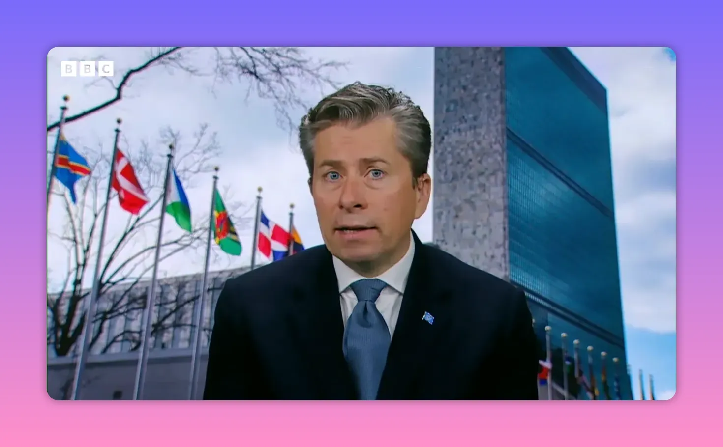 Intervenant en costume devant le siège des Nations unies avec des drapeaux en arrière‑plan, regard direct vers la caméra