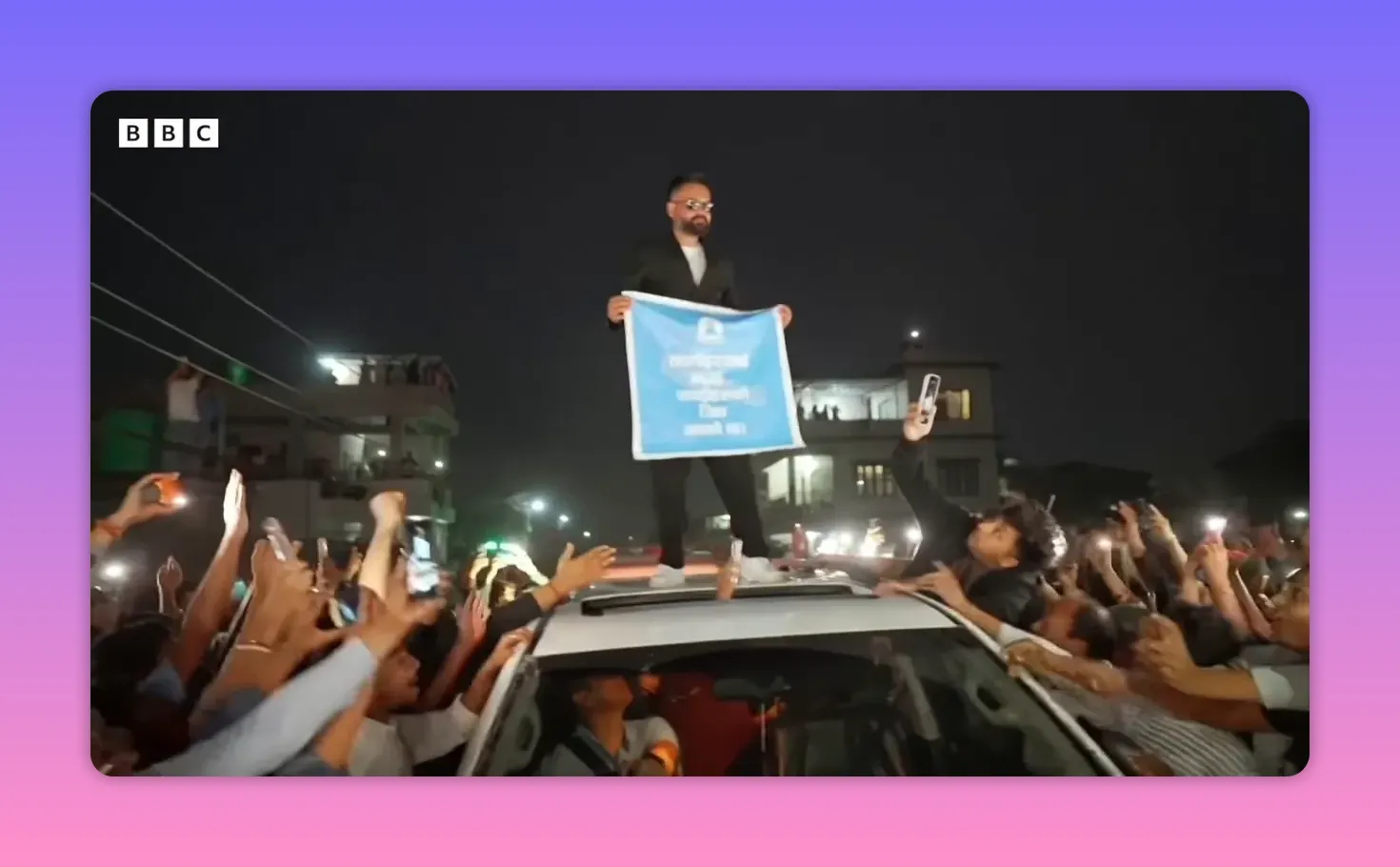 Homme en costume sombre et lunettes debout sur le toit d'une voiture tenant une banderole, entouré d'une foule la nuit
