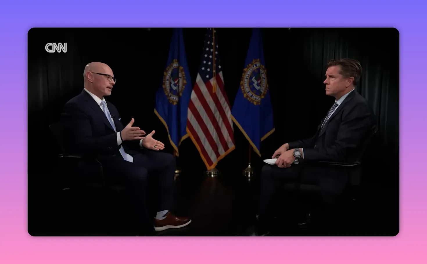 Zwei Personen in einem formellen Interview, US-Flaggen und Embleme im Hintergrund