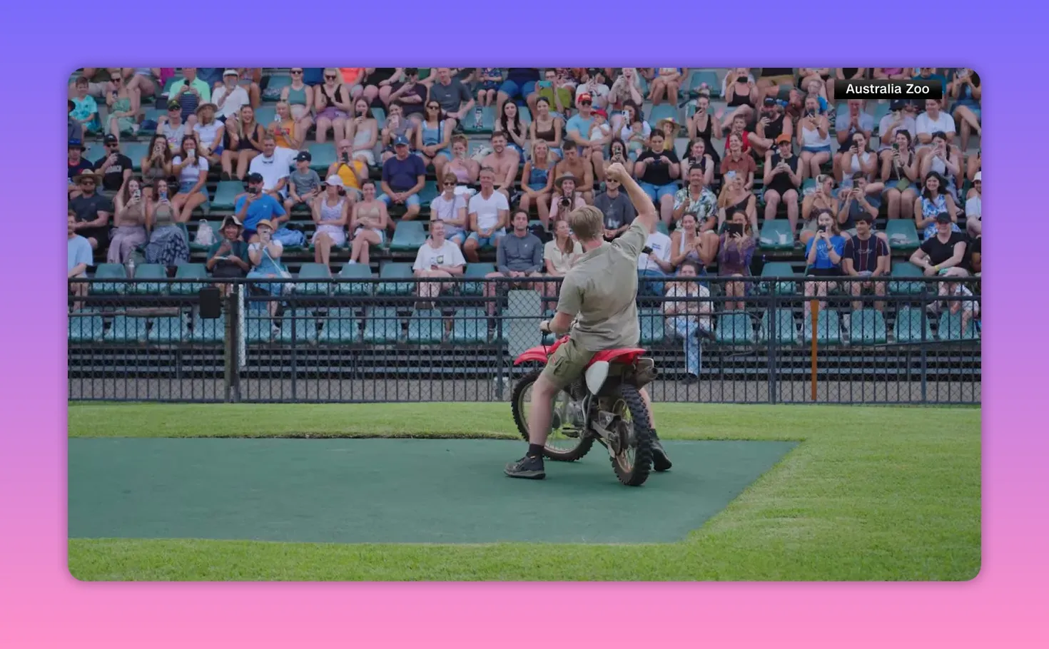 Rückansicht einer Person auf einem roten Motorrad, die dem jubelnden Publikum zuwinkt; große Tribüne im Hintergrund.