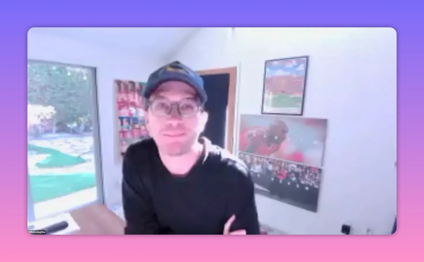 Bright, centered video screenshot of a presenter in a home office wearing a cap and glasses, looking at the camera with wall art behind them.