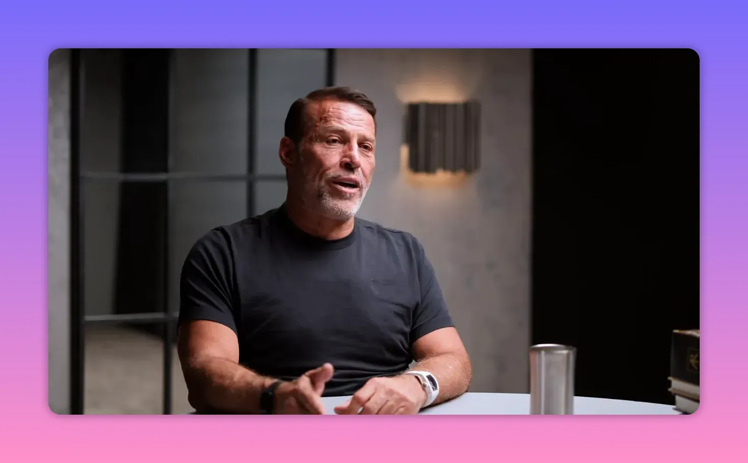 Speaker seated at a table explaining with one hand extended — clear, well-lit conversational portrait.