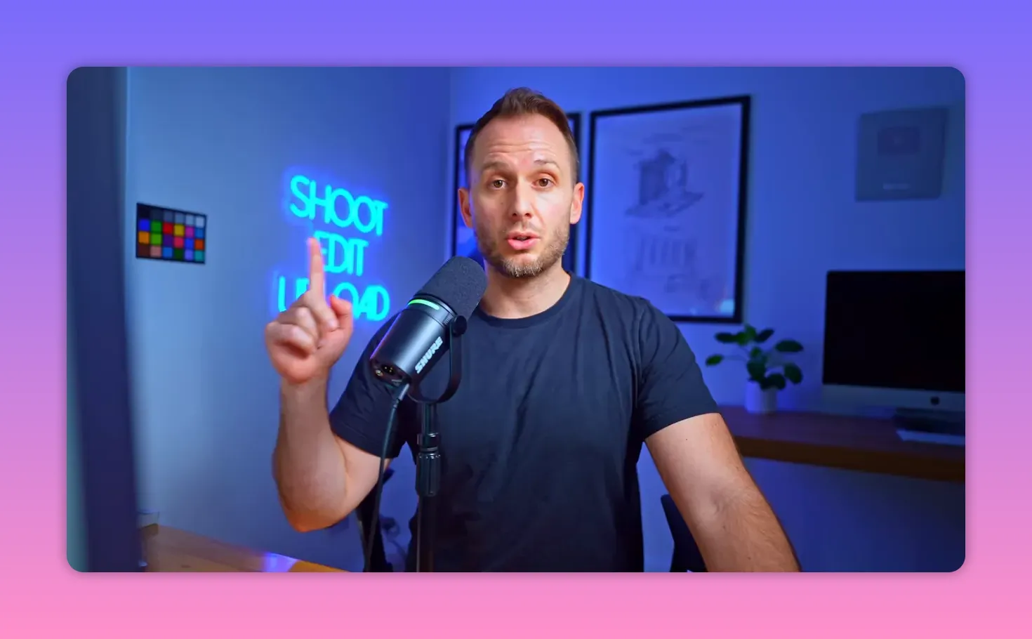 Presenter with one finger raised in a pointing/holding-up gesture beside a desk microphone and neon 'SHOOT EDIT UPLOAD' sign