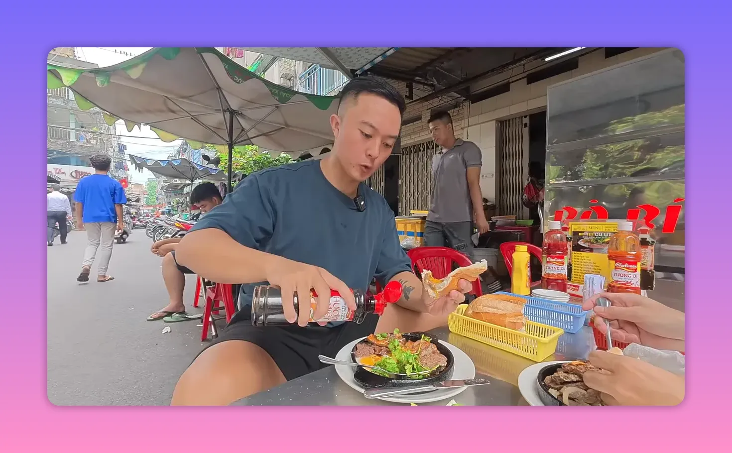 Person pouring sauce over a sizzling plate at a busy Vietnamese sidewalk food stall with diners, umbrellas and motorbikes in the background