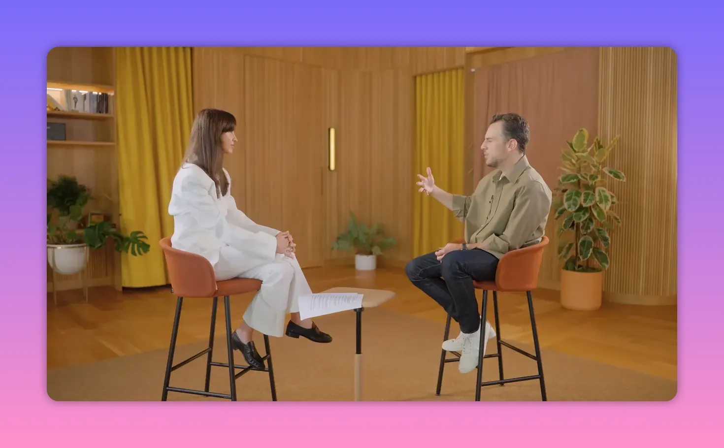 Wide interview frame of two people seated on stools in a warm, wood-paneled studio, gesturing while talking.
