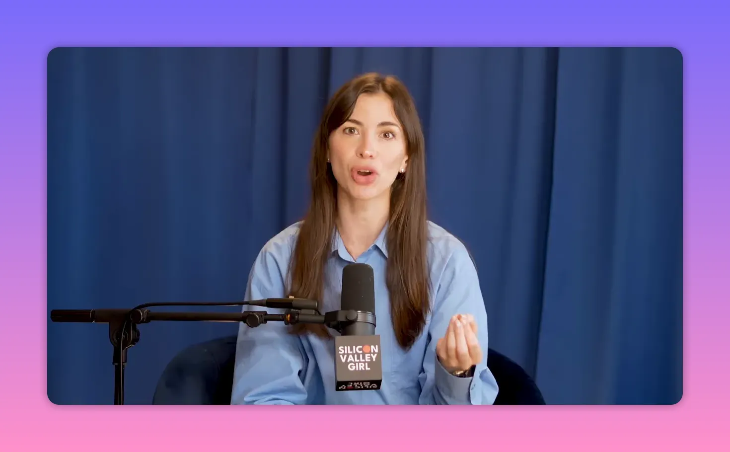 Silicon Valley Girl speaking into a microphone, gesturing with her hand, blue curtain background