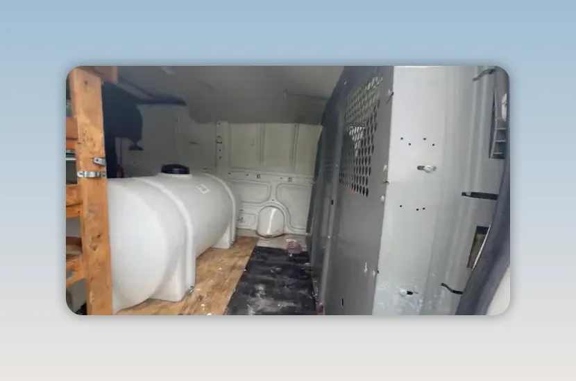 Cargo van interior with a large white water tank, wooden shelves, plywood floor, and metal partition