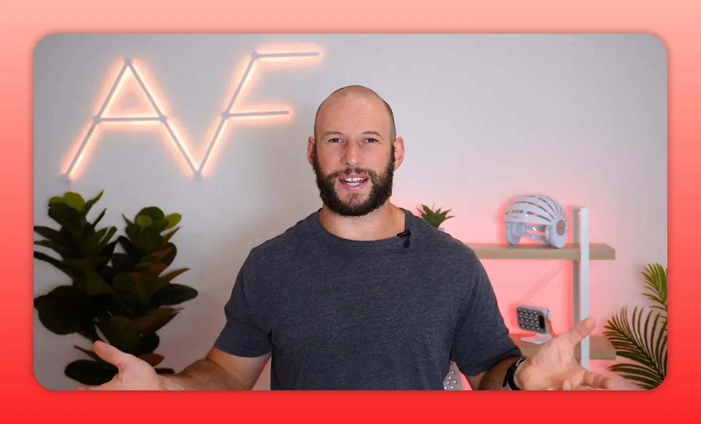 Presenter facing the camera with hands open in a welcoming gesture, neon AF sign and red-light therapy devices on a shelf behind him.