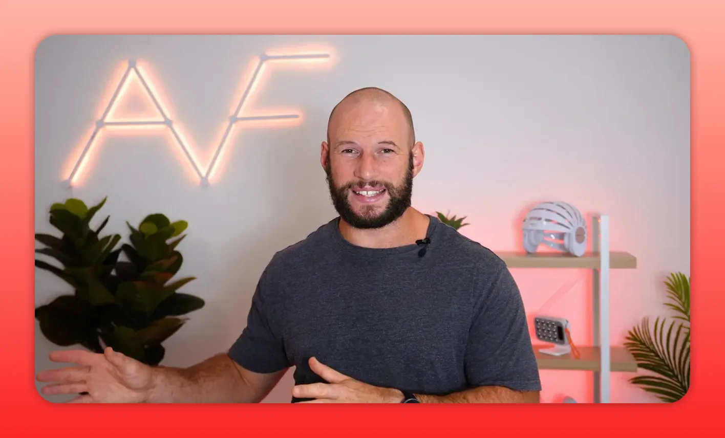 Presenter smiling and gesturing with hands visible, neon AF sign and red-light devices on shelving behind; clear, high-quality medium shot.