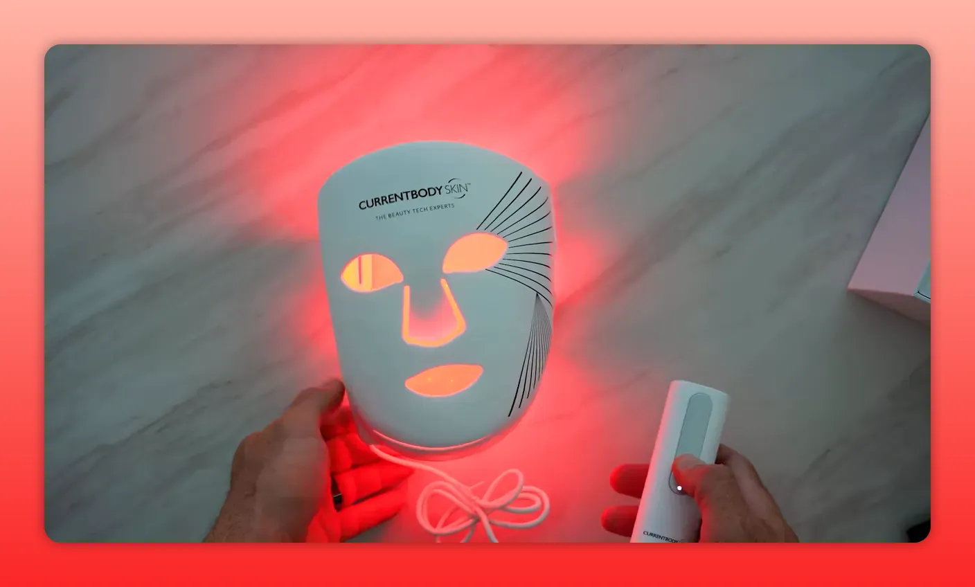 CurrentBody face LED mask glowing red with handheld controller and hands for scale