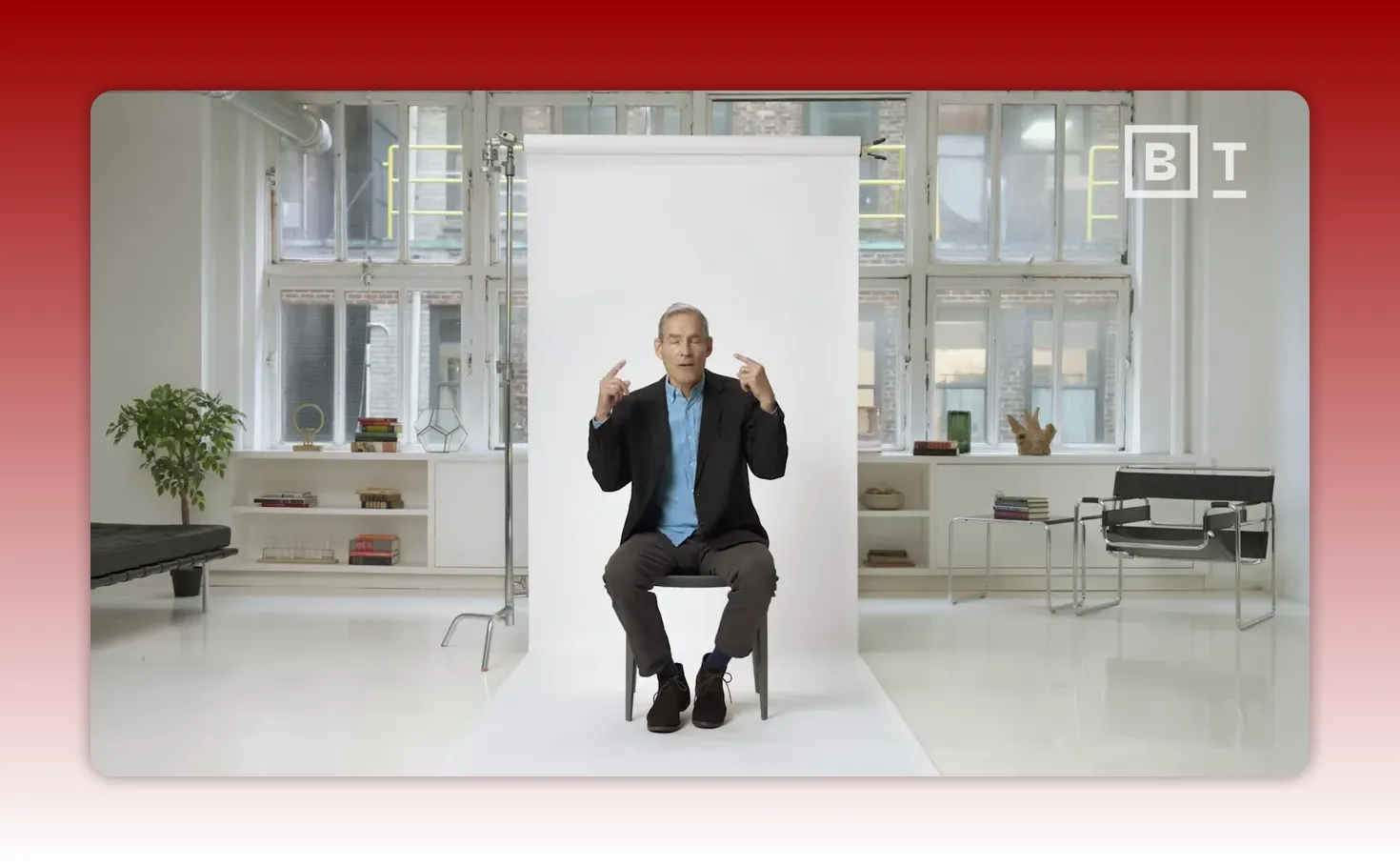 Speaker seated against a white backdrop pointing to his head with both index fingers to indicate thinking and attention.