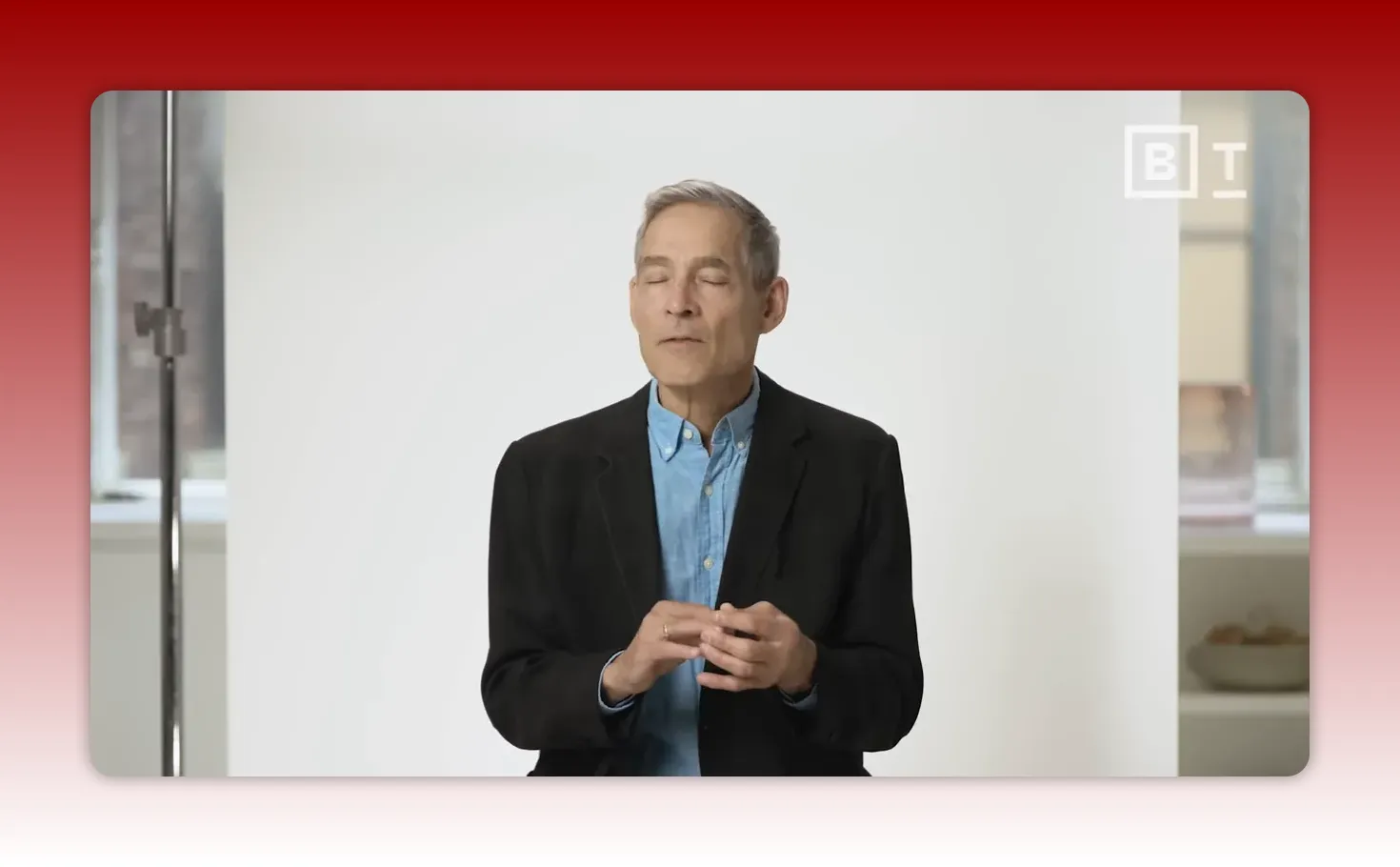 mid-shot of speaker with eyes closed and hands lightly clasped against a plain white studio backdrop, conveying reflection