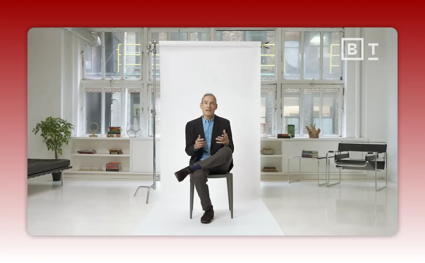 centered wide shot of a presenter speaking in a white studio with a backdrop, chair, and light furniture visible