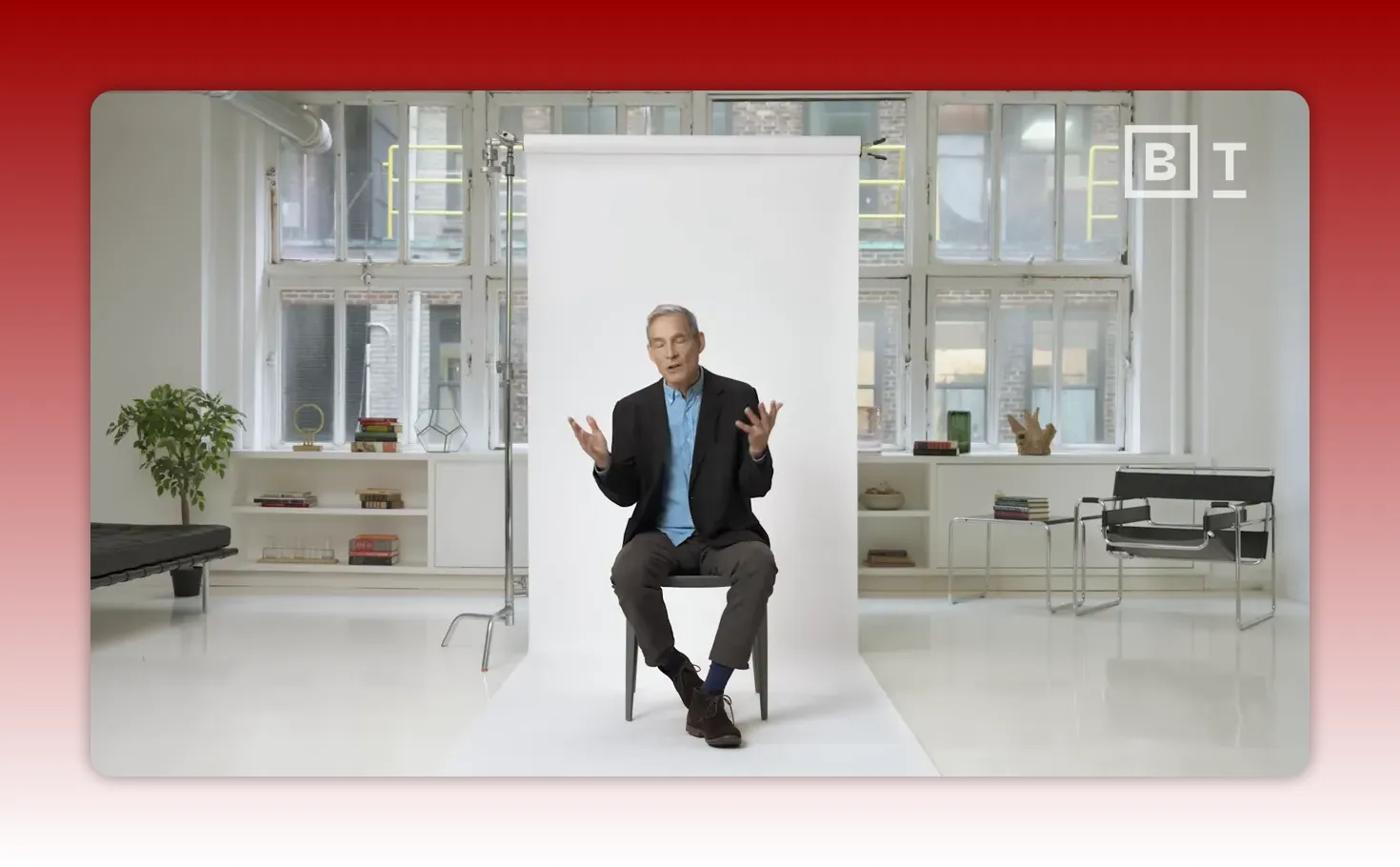 Full-body studio shot of the speaker seated with open-hand gestures in front of a white backdrop