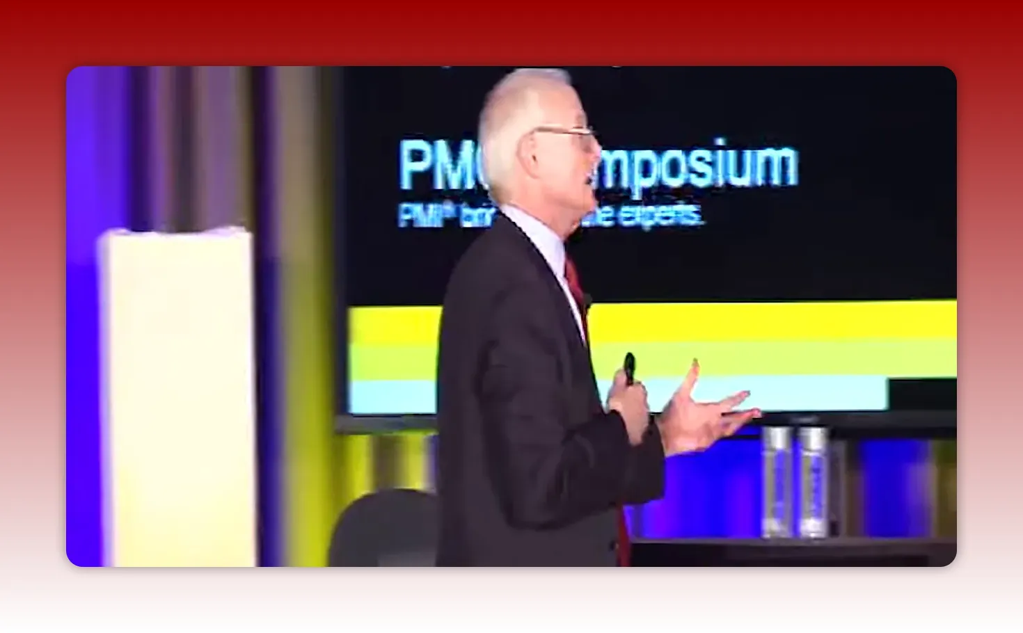 Presenter in side profile gesturing with a visible 'PMO Symposium' slide and horizontal colored bars behind him