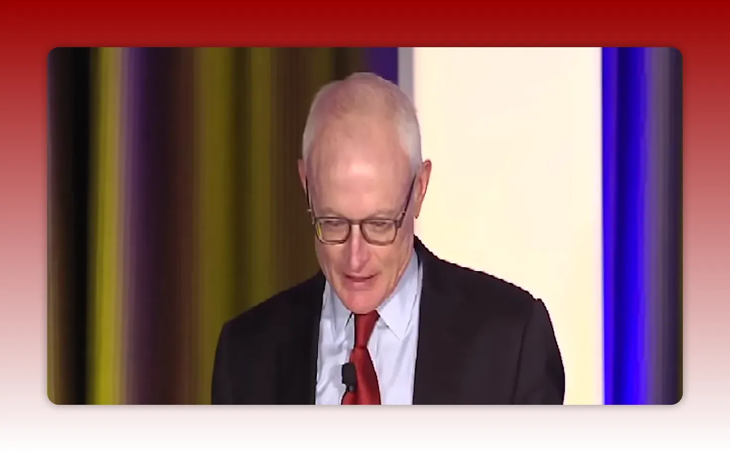 Presenter in suit and red tie with lavalier microphone on stage, clear front-facing view