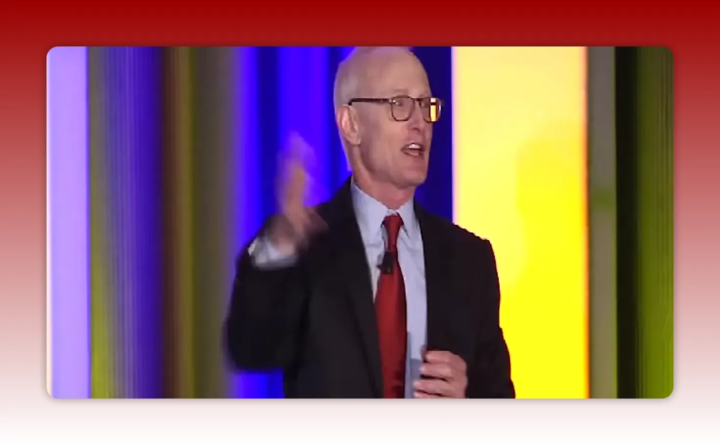 Presenter in suit and red tie gesturing with hand while speaking on stage against a colorful vertical backdrop, clear frontal view