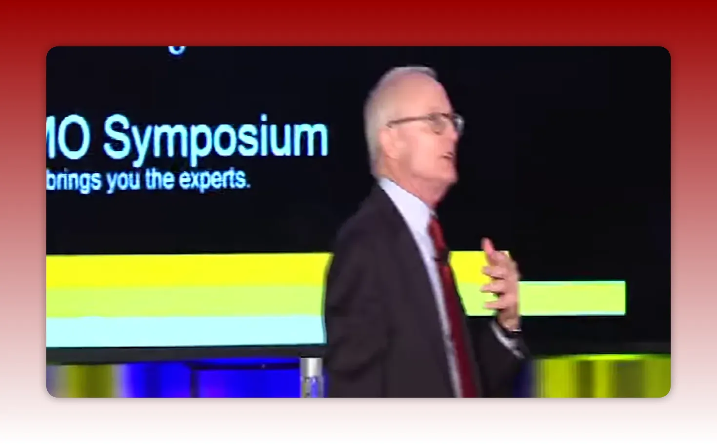 Presenter at the edge of the stage next to a slide that reads 'PMO Symposium' with horizontal colored bars — illustrates project-management context