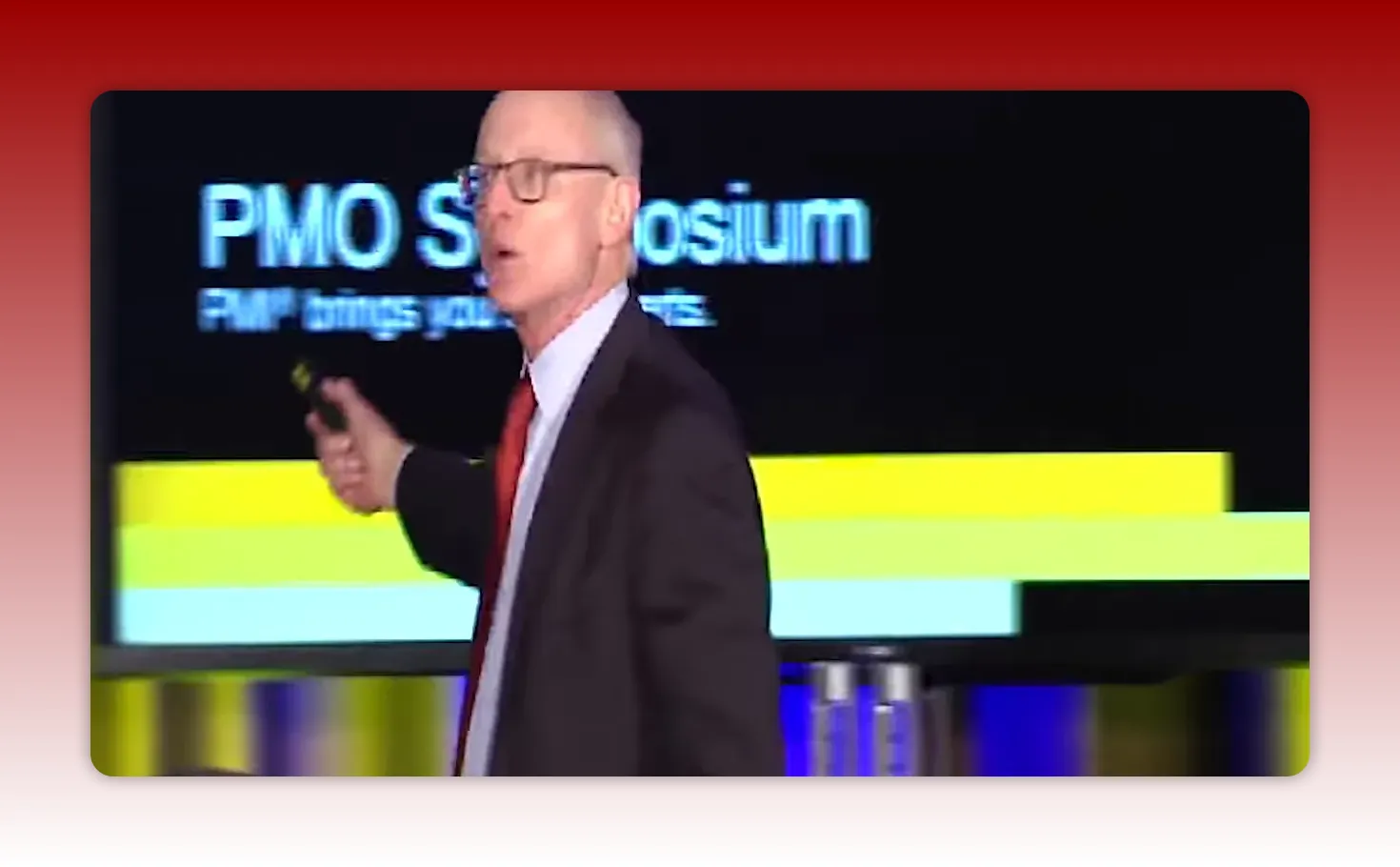 Presenter on stage pointing toward a slide that reads 'PMO Symposium' with horizontal colored bars, illustrating project management context